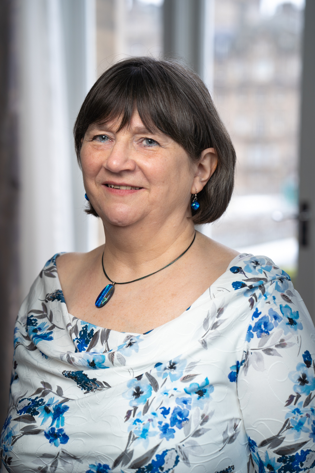 A woman with short, dark hair wearing a white blouse with blue floral patterns, a blue pendant necklace, and matching blue earrings.