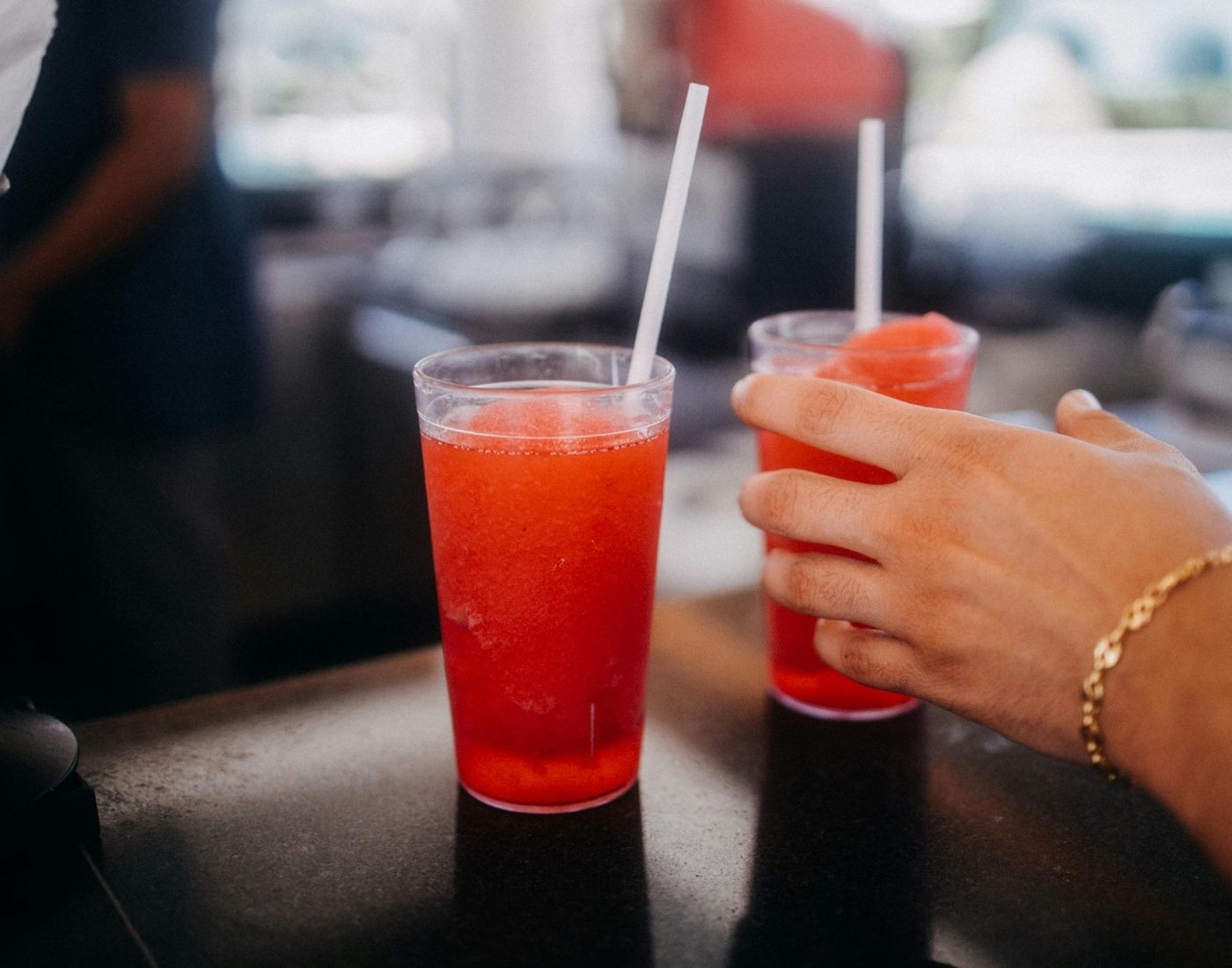 Two plastic cups filled with red slushy drinks, each with a white straw. One cup is held by a hand wearing a gold bracelet. The background is an indoor setting with blurred objects and people.