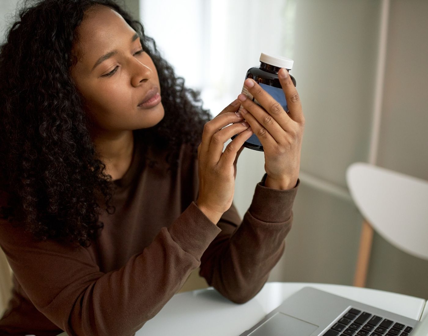 A person examines a medication bottle closely while sitting at a desk with a laptop.
